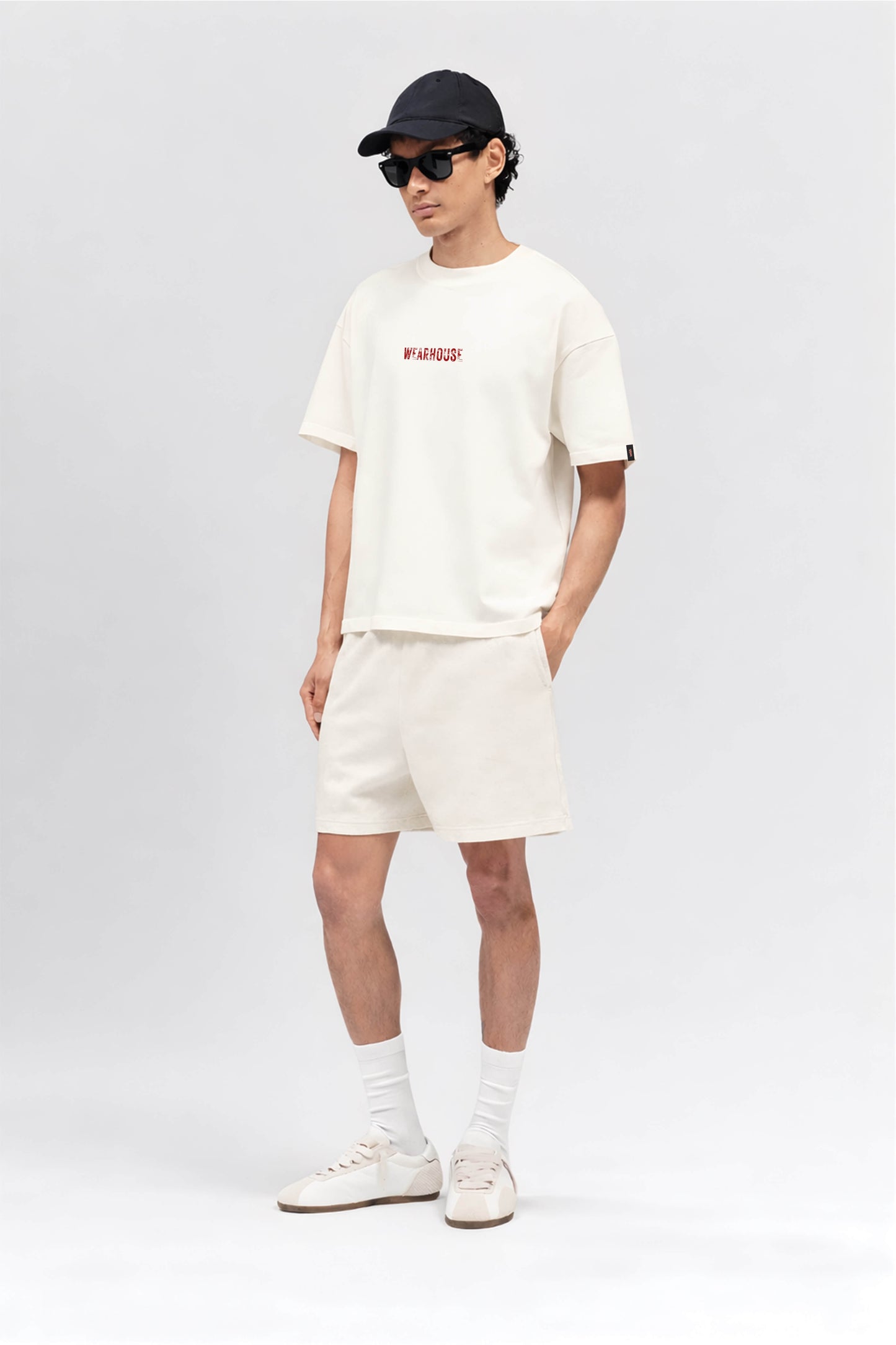 Person wearing an off white t-shirt and shorts set with a black cap and sunglasses on a light gray background