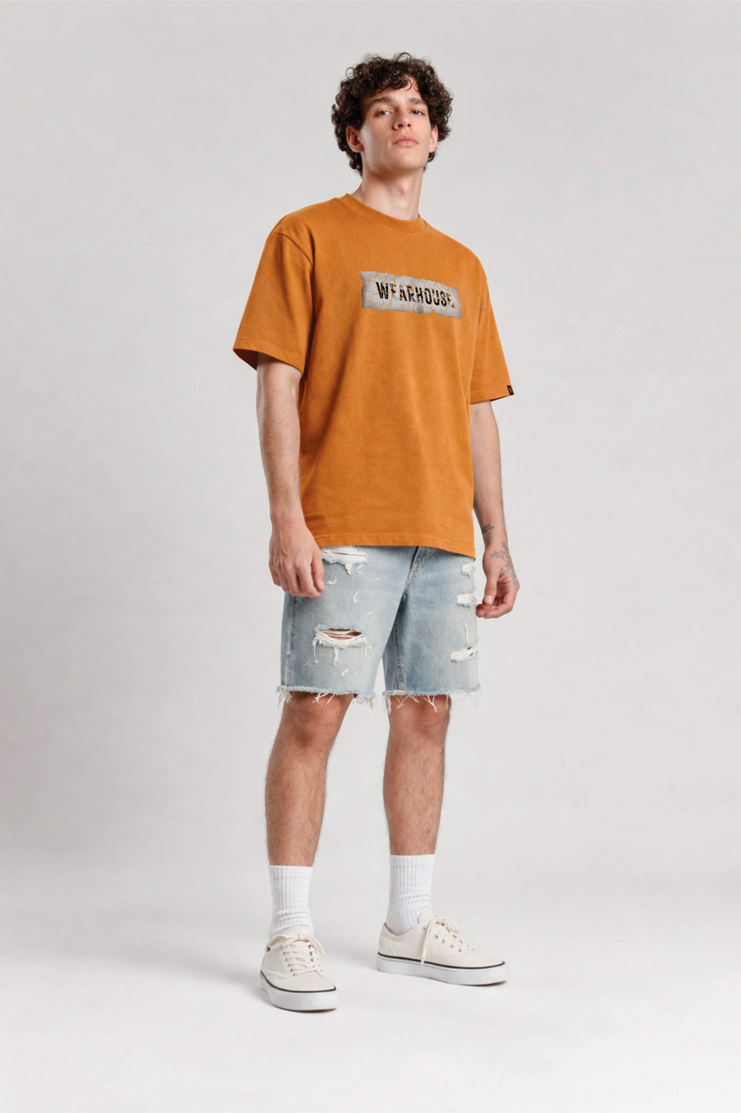 Man wearing an orange t-shirt with a logo and light blue denim shorts on a white background