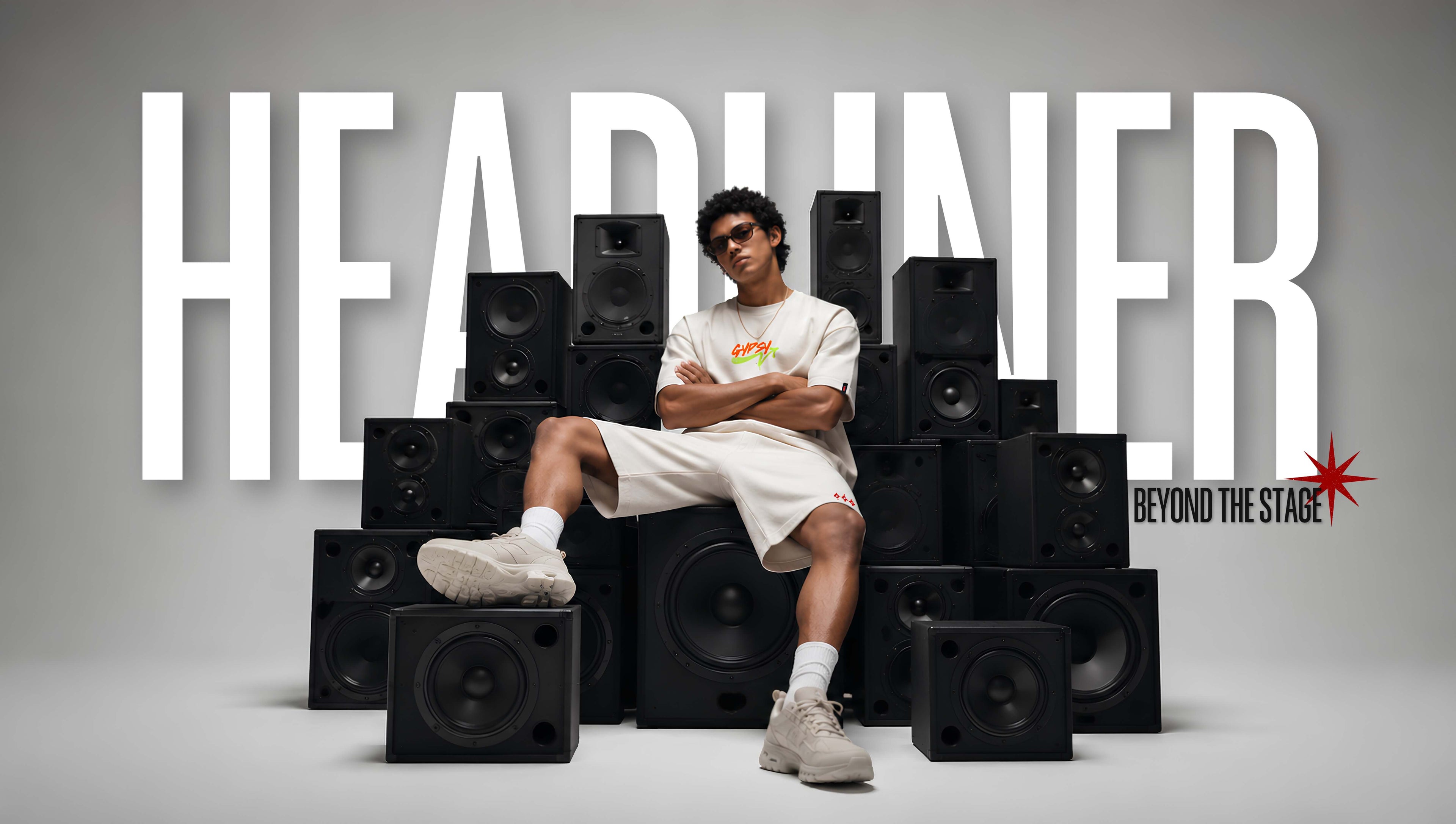A man wearing a white graphic t-shirt, white shorts, and white sneakers sits cross-legged among a large stack of black speakers. The background is a clean gray studio setting with the word 'HEADUNER' in large white letters and 'BEYOND THE STAGE' with a red star logo in the bottom right.