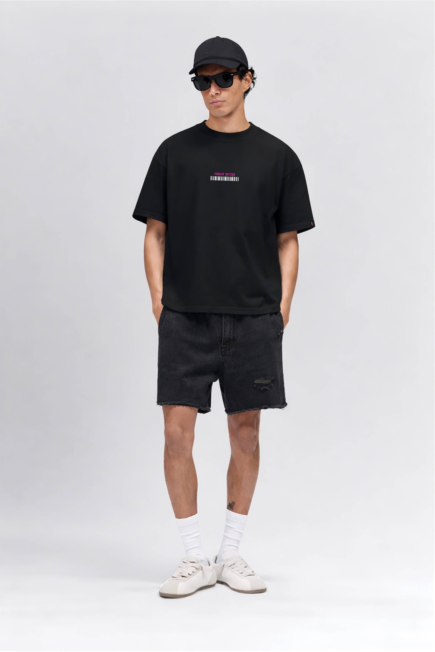 Man wearing a black t-shirt with a logo, black shorts, white sneakers, and a black cap on a white background