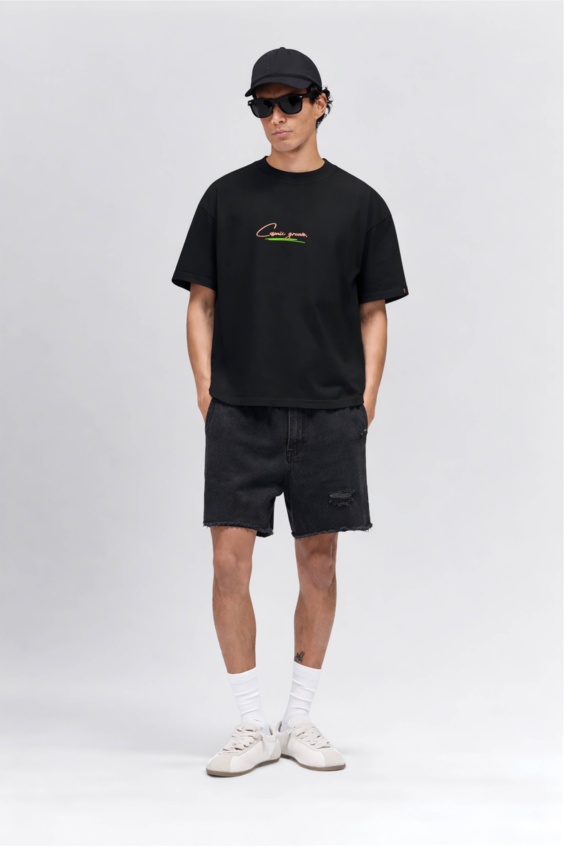Man wearing a black t-shirt with a logo, black shorts, white sneakers, and a black cap on a white background