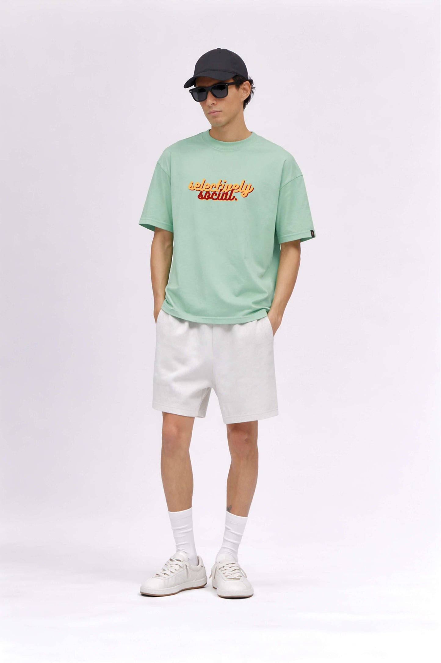Person wearing a light green t-shirt with text, white shorts, white sneakers, and a black cap on a white background