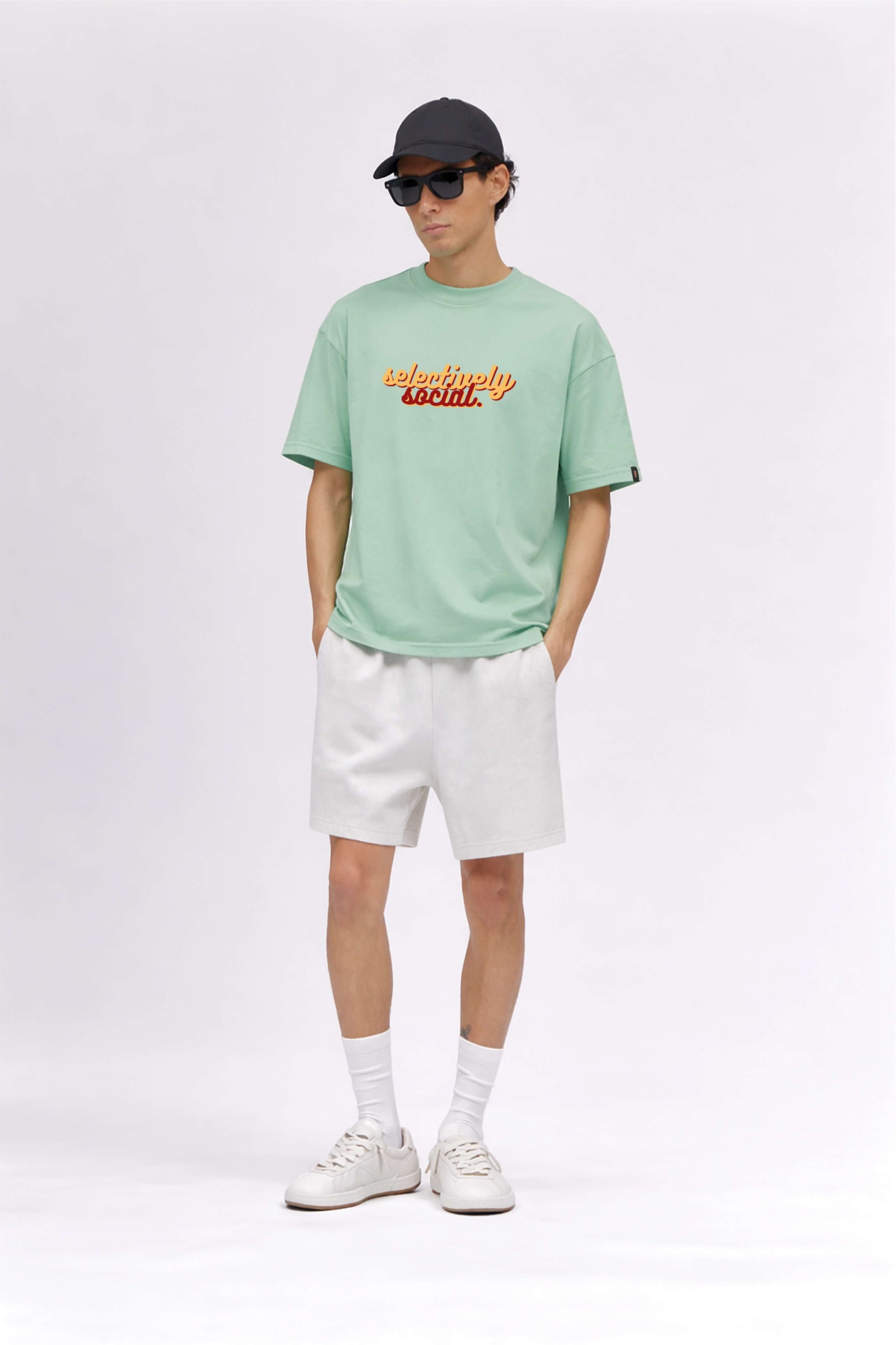 Person wearing a light green t-shirt with text, white shorts, white sneakers, and a black cap on a white background