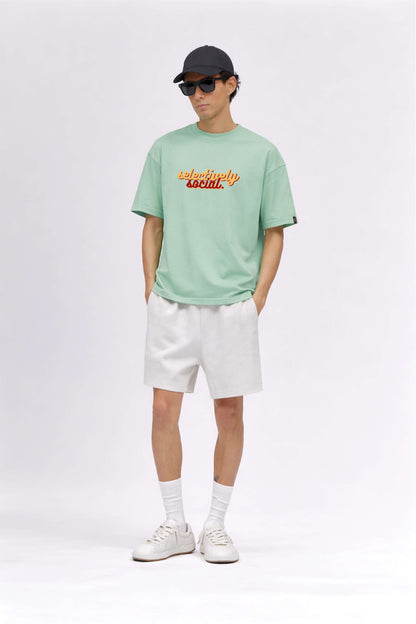 Person wearing a light green t-shirt with text, white shorts, white sneakers, and a black cap on a white background