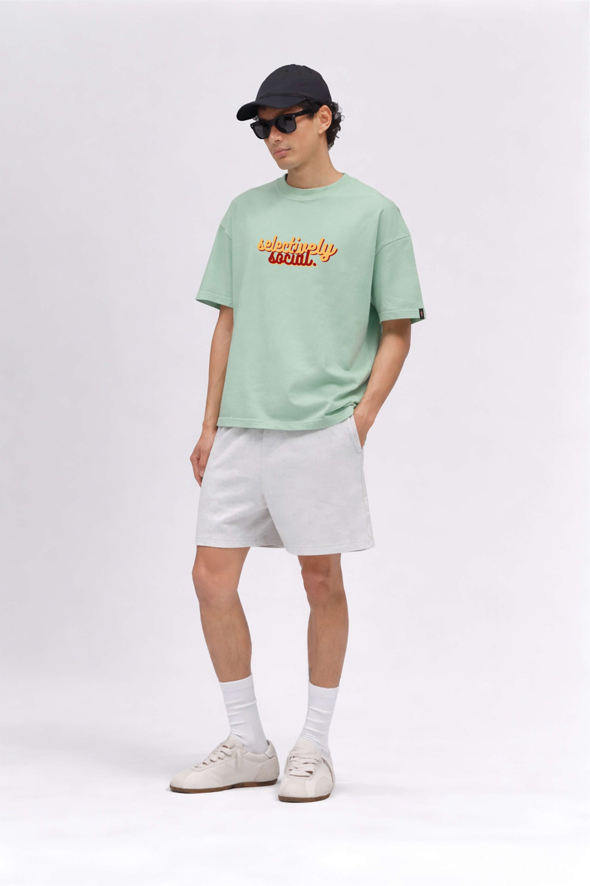Person wearing a light green t-shirt with text, white shorts, and a black cap on a white background