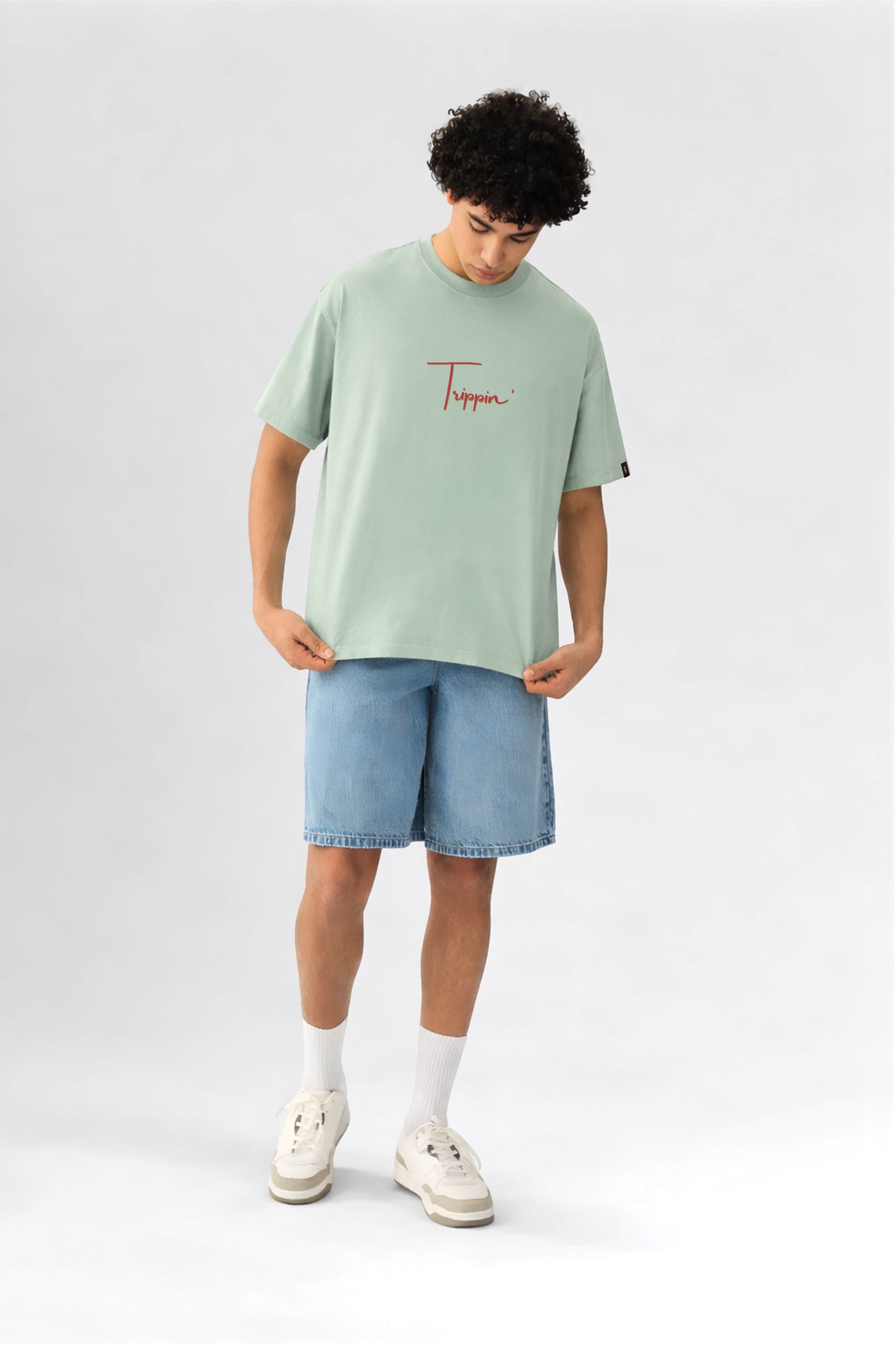 Person wearing a light green t-shirt with a logo and blue shorts on a white background