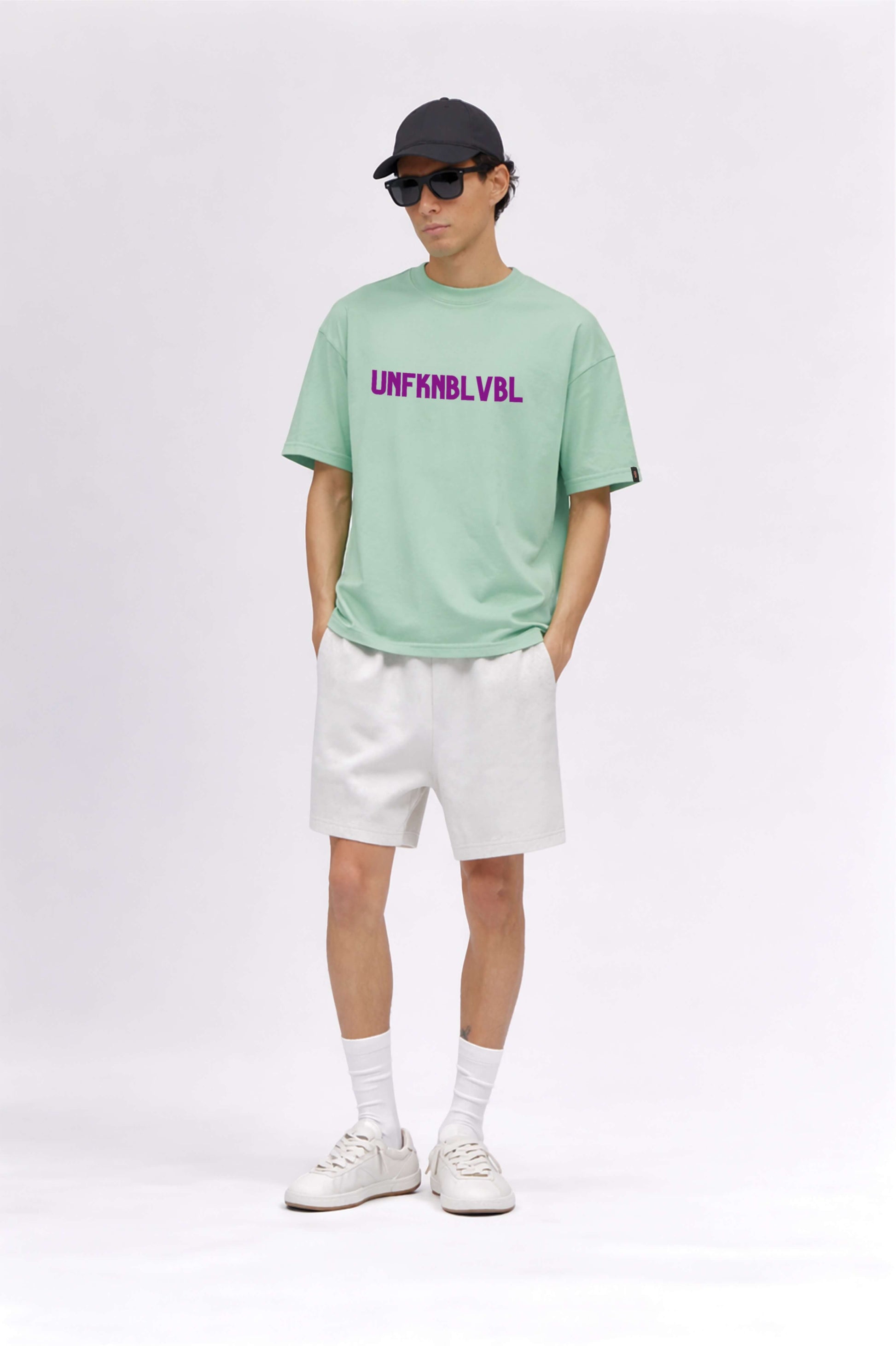 Person wearing a light green t-shirt with text, white shorts, white sneakers, and a black cap on a white background