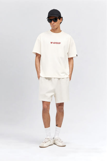 Person wearing an off white t-shirt with red text and white shorts on a light gray background