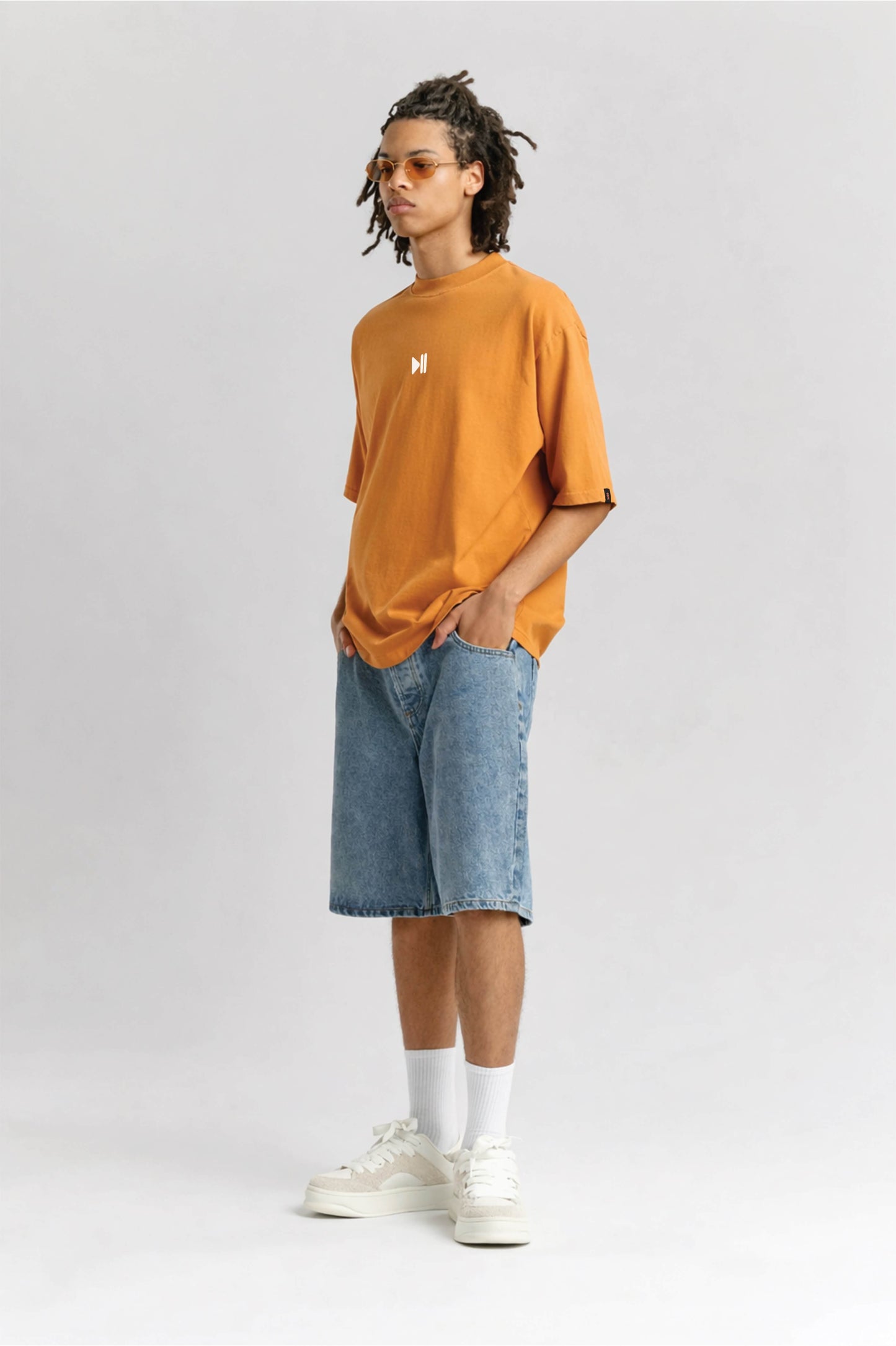 Person wearing an orange t-shirt with a logo, blue shorts, and white sneakers on a light gray background