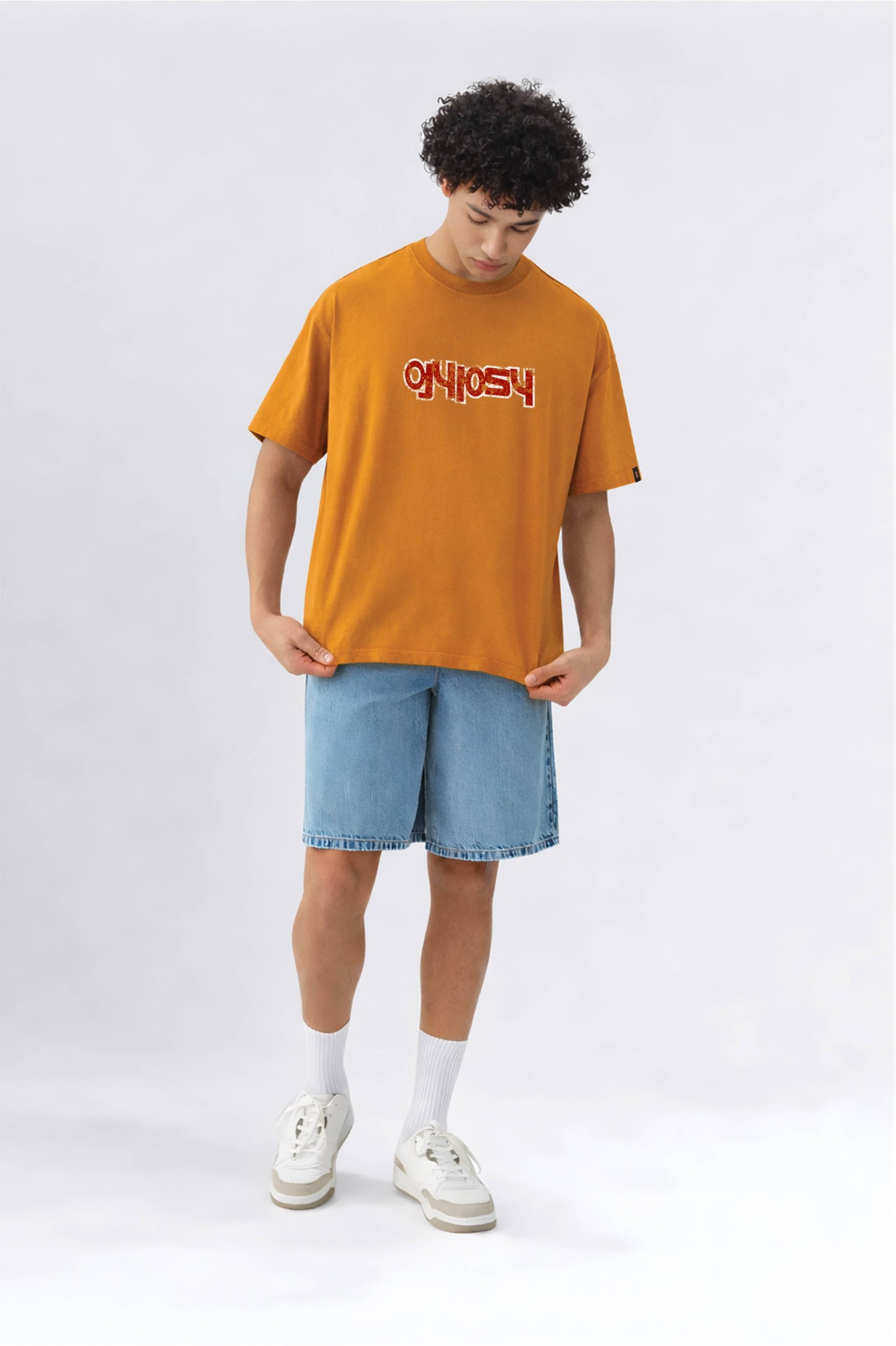Person wearing an orange t-shirt with text and light blue shorts on a white background
