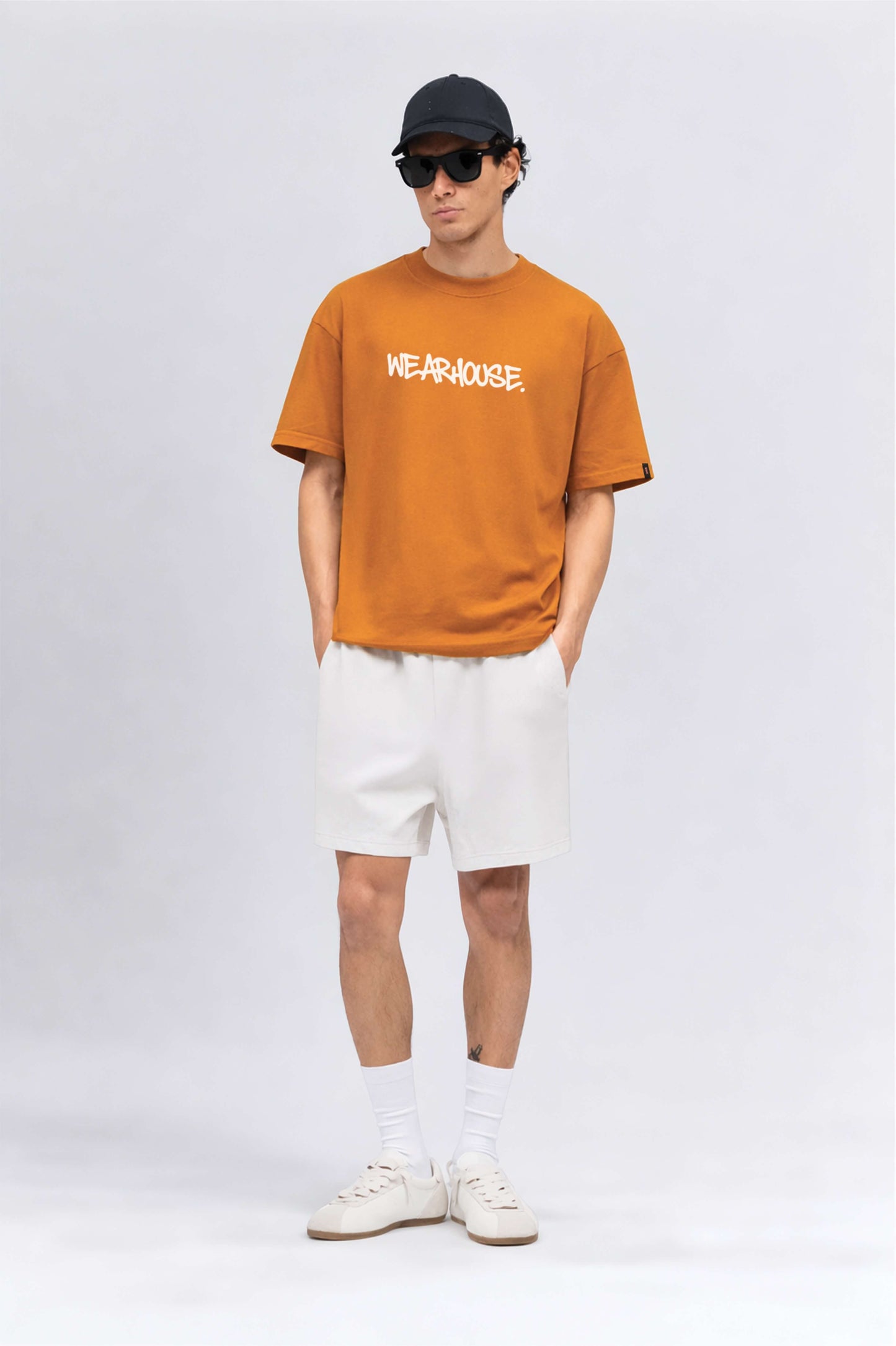 Person wearing an orange t-shirt with text, white shorts, and a cap on a plain background