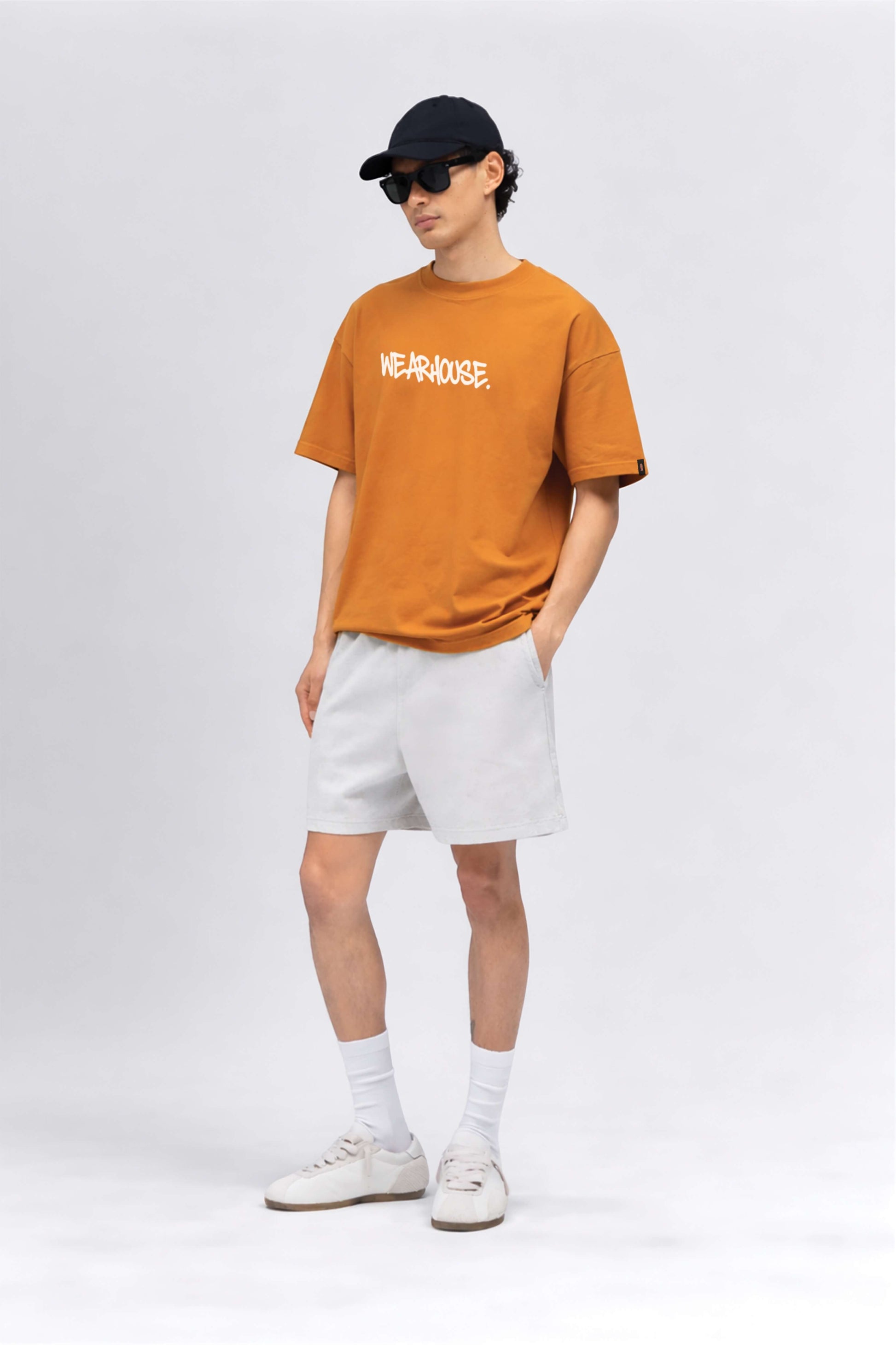 Person wearing an orange t-shirt with text, white shorts, and a black cap on a white background